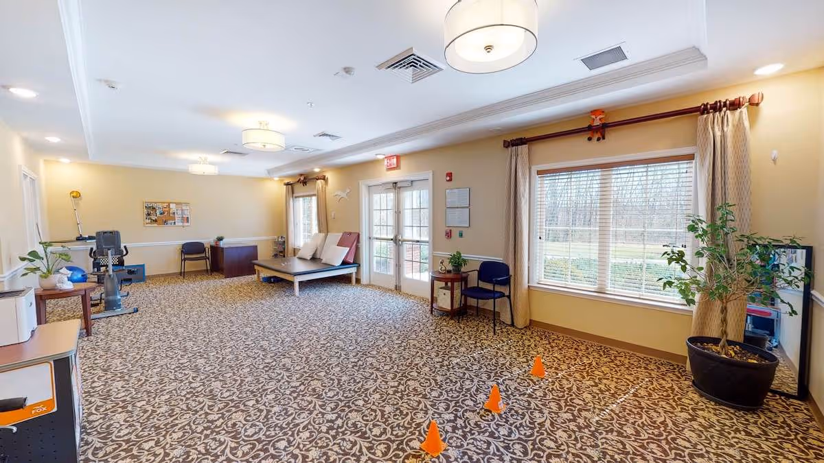 A spacious room with patterned carpet, beige walls, and ceiling lights. The room contains exercise equipment, a padded bench with pillows, chairs, a desk, and several plants. Large windows with curtains allow natural light to enter, and there are double glass doors leading outside. Orange cones are placed on the floor near the windows.