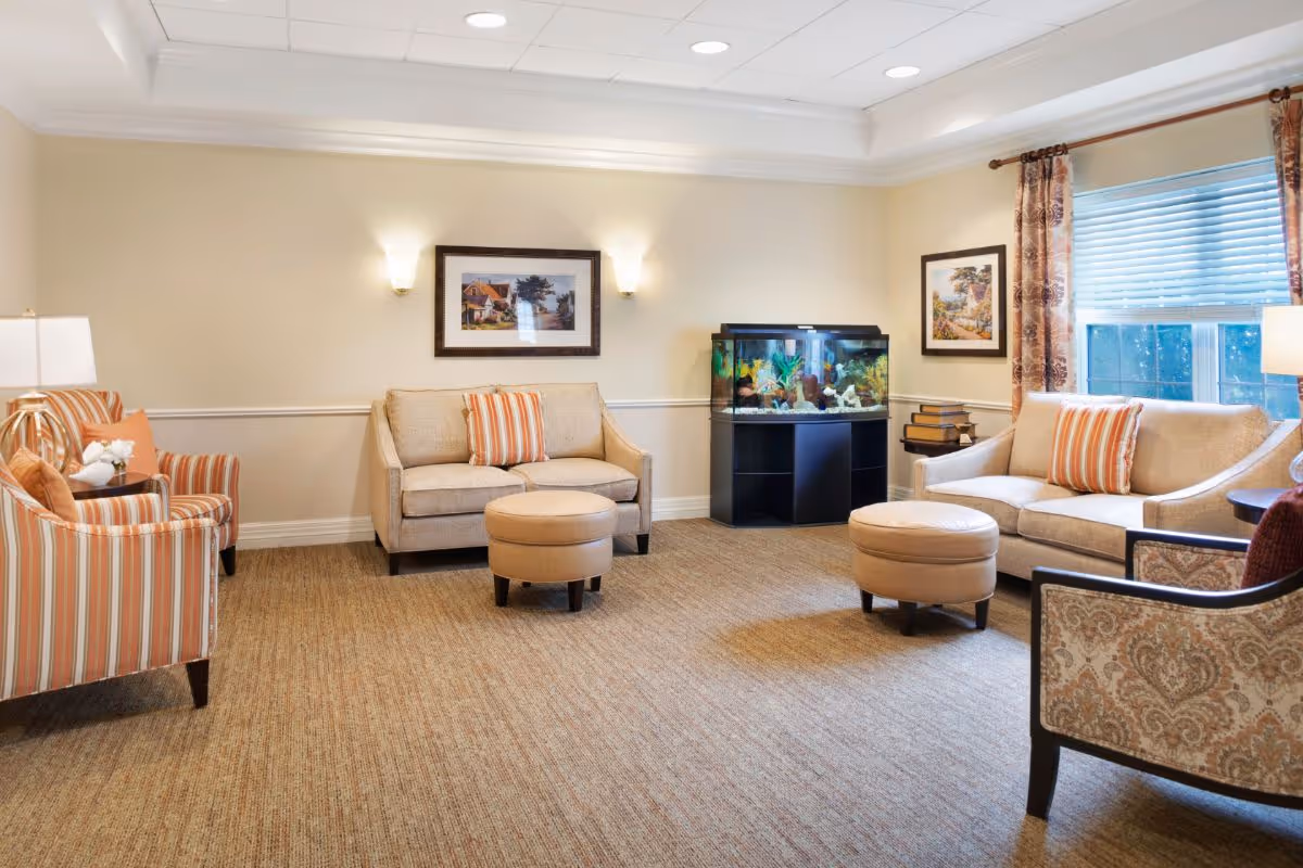 A cozy living room with beige walls and carpet, featuring two beige sofas with striped orange and beige pillows, two round beige ottomans, two striped armchairs, a patterned armchair, a fish tank on a black stand, framed artwork on the walls, and a window with patterned curtains letting in natural light.