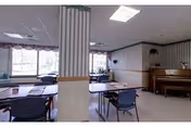 Bright common room with several tables and chairs, a piano against the wall, large windows, and a central striped column.