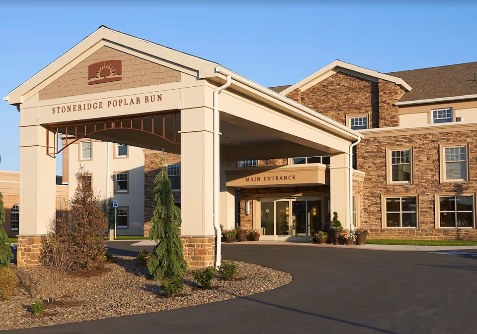 Exterior view of the main entrance of Stoneridge Poplar Run, a retirement living community. The building features a covered driveway with columns and a sign displaying the community name. The facade is made of stone and beige siding with multiple windows. There are landscaped areas with small trees and shrubs around the entrance.