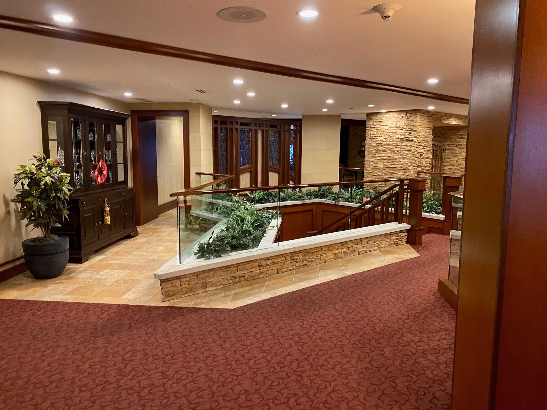 Interior view of a senior living facility hallway with a red patterned carpet, a glass railing enclosing a planter with green plants, a dark wooden cabinet with glass doors on the left, and stone accent walls. The area is well-lit with recessed ceiling lights.