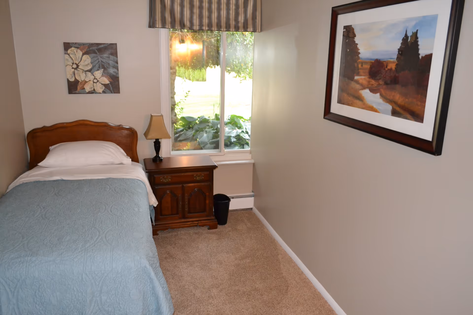 A small bedroom with a single wooden bed covered with a light blue quilt and white pillow. Next to the bed is a wooden nightstand with a lamp on it. There is a window with a striped valance showing greenery outside. The walls are beige, decorated with a floral painting above the bed and a landscape painting on the adjacent wall. The floor is carpeted in a light brown color.