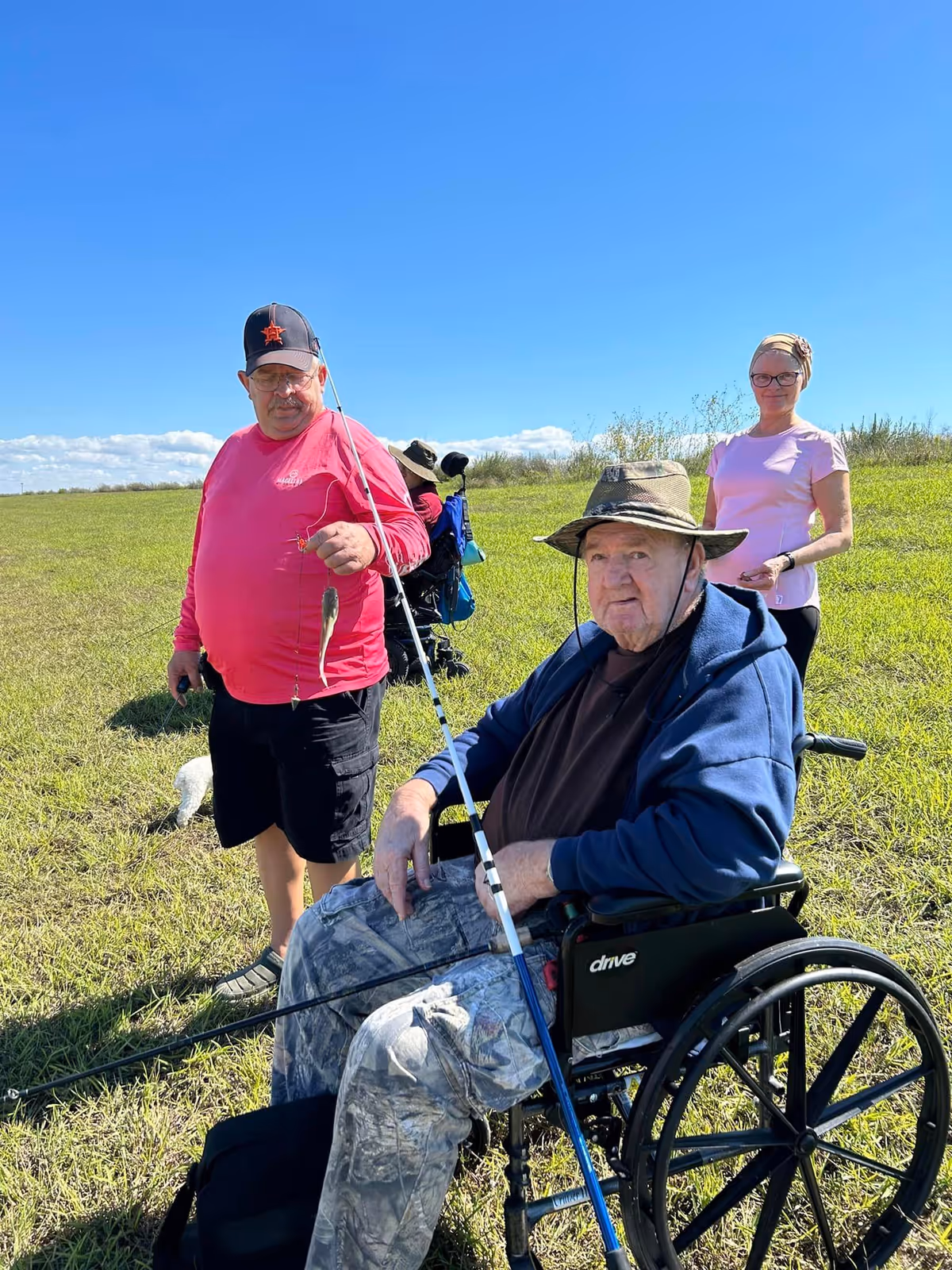 Three people outdoors on a sunny day in a grassy field. One man in a wheelchair wearing a hat and blue jacket holds a fishing rod. Another man in a red long-sleeve shirt and black shorts stands next to him holding a fishing line with a small fish. A woman in a pink shirt and glasses stands behind them smiling. The sky is clear and blue.