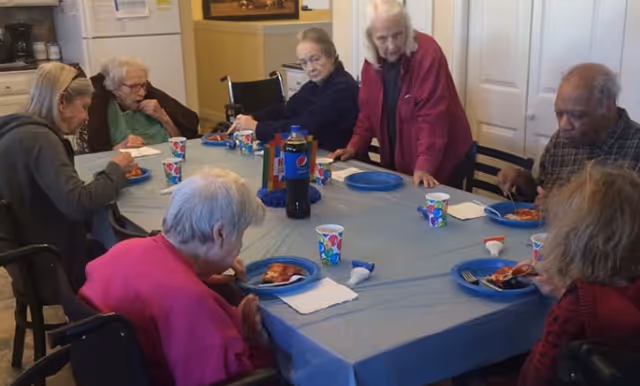 A group of elderly people sitting around a table covered with a blue tablecloth, eating pizza and drinking from colorful paper cups in a dining area.