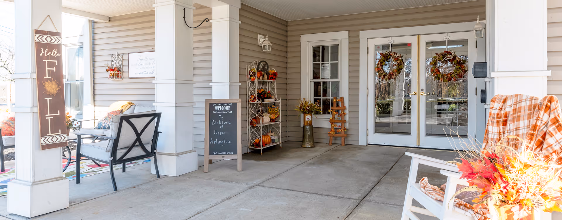 Covered outdoor entrance area of Bickford of Upper Arlington decorated for fall with a 'Hello Fall' sign, a welcome chalkboard, wreaths on glass double doors, chairs with cushions and blankets, and autumn-themed floral arrangements.