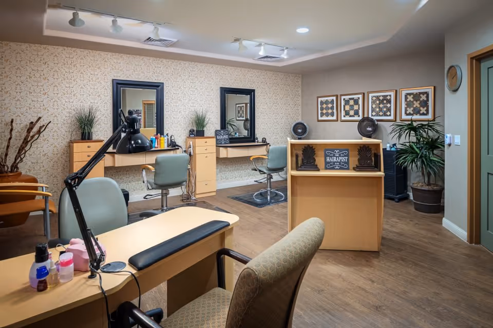 Interior salon area with styling stations, chairs, mirrors, and manicure tables in a senior living facility.