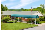 Exterior view of a single-story brick building with a green awning over the entrance and an American flag on a flagpole in front, surrounded by green grass and shrubs.