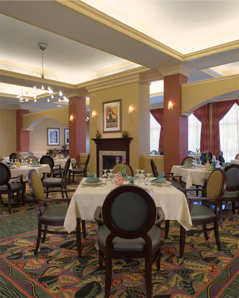 Elegant dining room with round tables covered in white tablecloths, set with plates, glasses, and folded napkins. The room features patterned carpet, upholstered chairs, warm lighting from wall sconces and a modern chandelier, and large windows with red curtains allowing natural light.