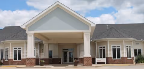 Front entrance of a single-story senior living facility with a covered porte-cochere, columns, brick accents, and double glass doors under a partly cloudy sky.