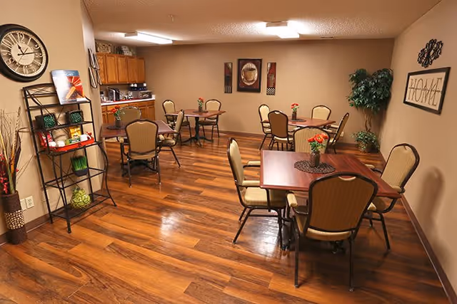 A cozy dining room with wooden floors and beige walls, featuring several square tables each surrounded by four cushioned chairs. Each table has a small flower arrangement. On the left side, there is a black metal shelving unit with decorative items and a large wall clock above it. The back wall has framed artwork, and a green potted plant is placed in the corner. The room is softly lit with ceiling lights.