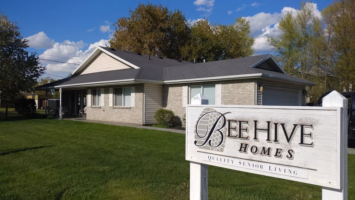 Single-story brick and siding building with a gray roof, surrounded by green grass and trees under a blue sky with clouds. In the foreground, there is a white wooden sign that reads 'Beehive Homes Quality Senior Living'.