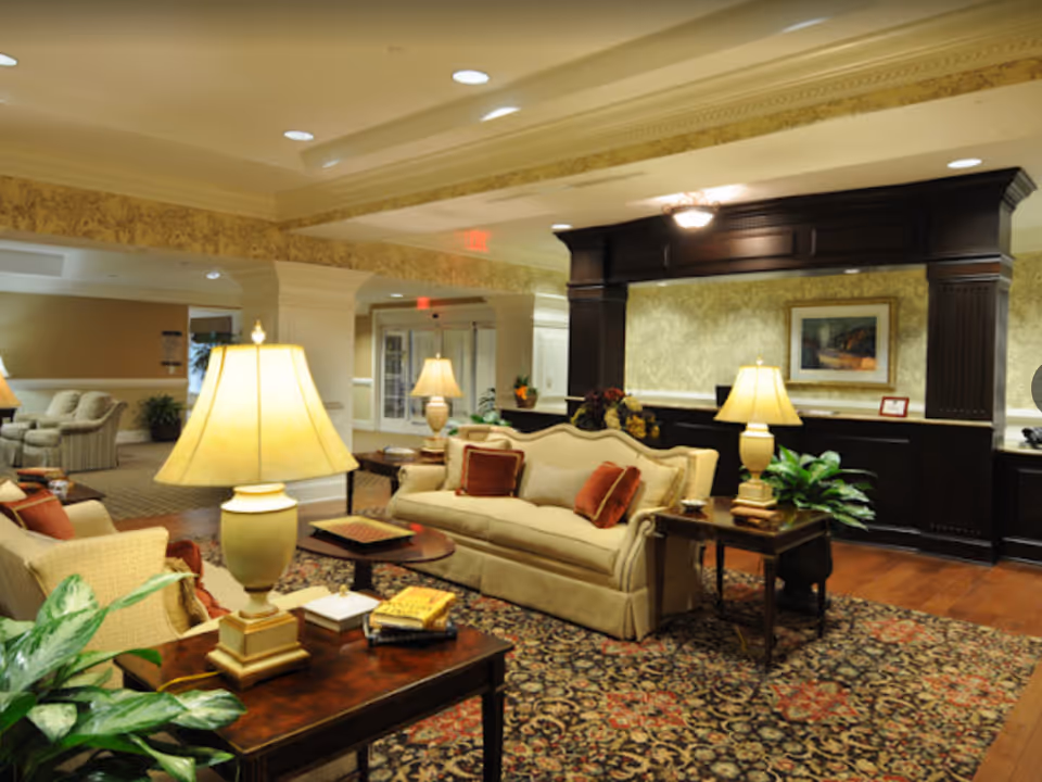 A warmly lit living room area in a senior living facility with beige sofas, wooden side tables with lamps, a patterned carpet, and a dark wood reception desk in the background. The walls have a light floral wallpaper and there are framed pictures and plants decorating the space.