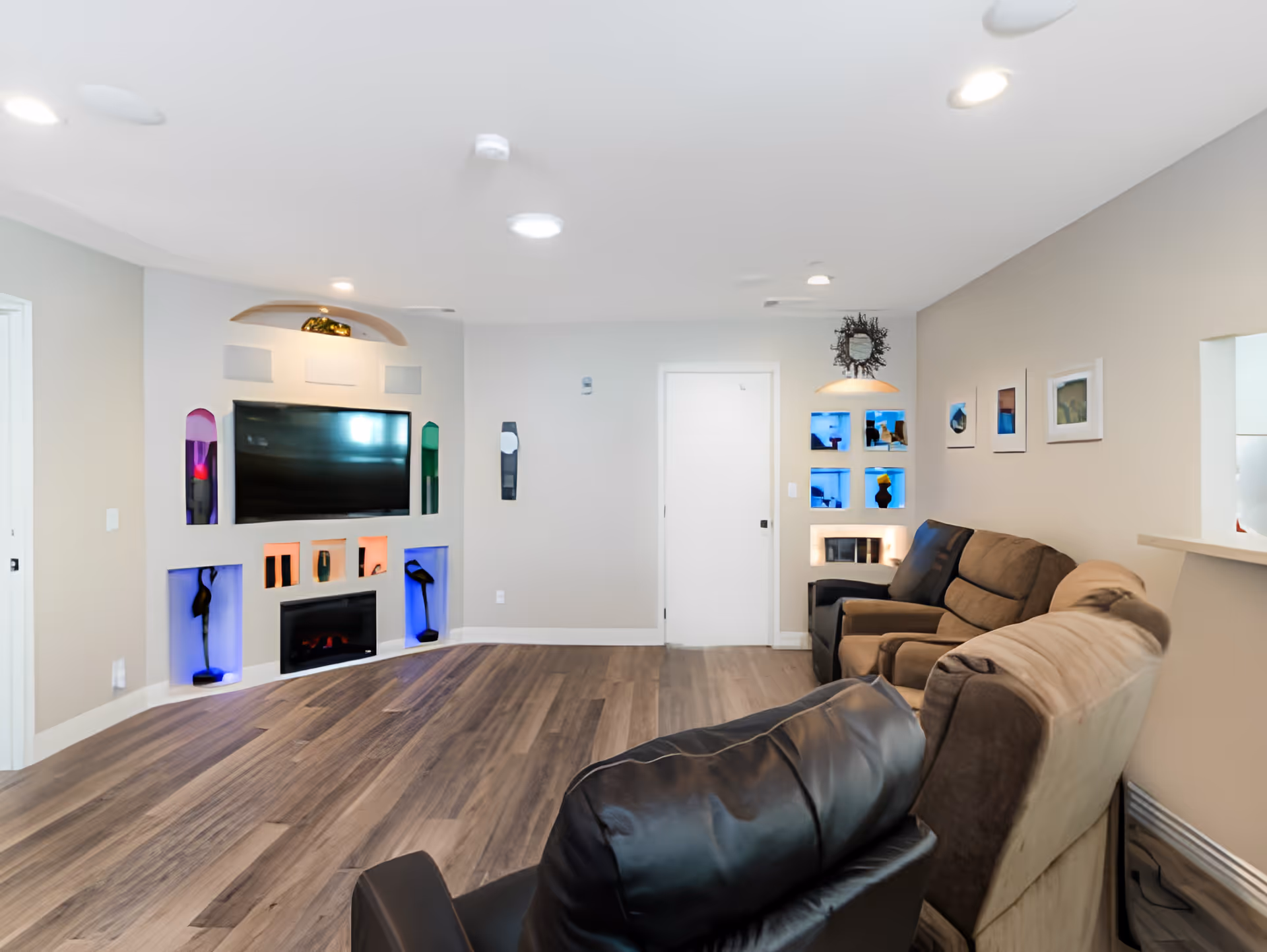 A modern living room with wood flooring, a wall-mounted flat-screen TV above a built-in electric fireplace, and decorative niches with colorful lighting. The room has a beige and light gray color scheme, with a black leather chair and a brown sofa facing the TV. There are framed pictures on the wall and a small window opening to an adjacent room.