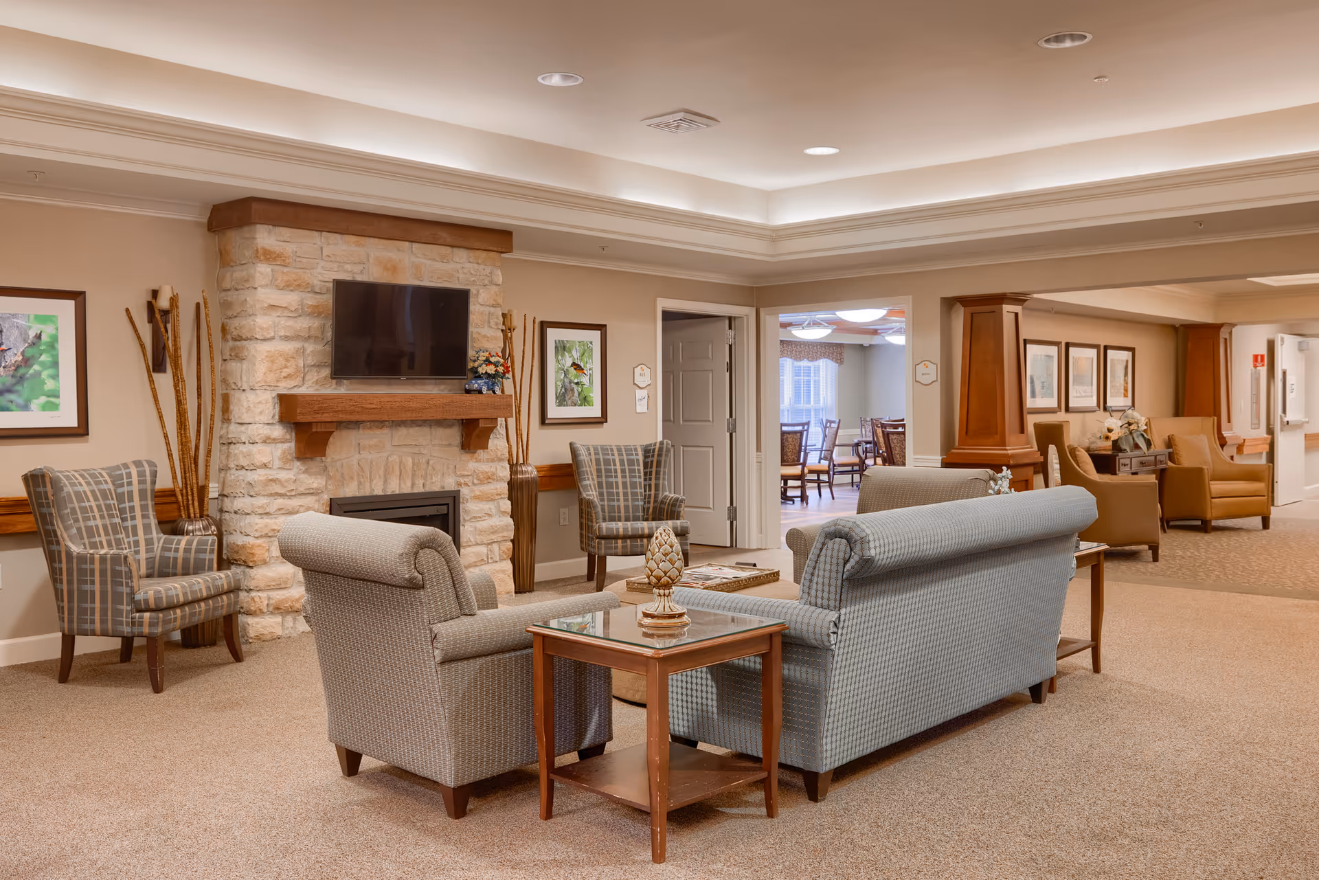 A cozy senior living facility common area with a stone fireplace and a mounted flat-screen TV. The room features comfortable upholstered armchairs and sofas arranged around a glass-top coffee table. The walls are decorated with framed artwork, and there is soft, warm lighting from recessed ceiling lights. In the background, there is a dining area with tables and chairs visible through an open doorway.