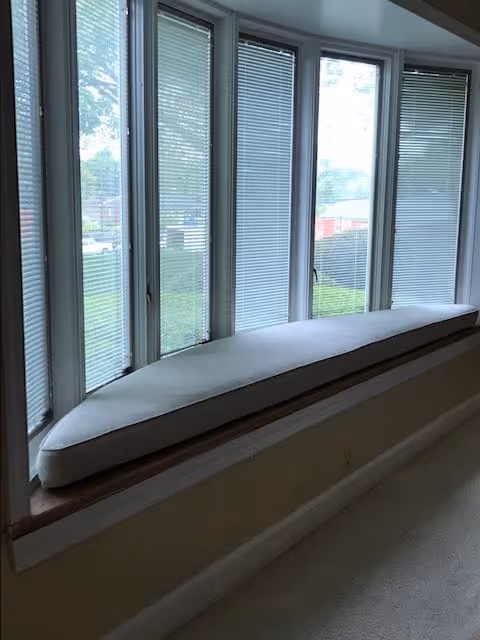 A curved window seat with a light gray cushion positioned in front of a series of tall windows with closed blinds, overlooking a grassy outdoor area.