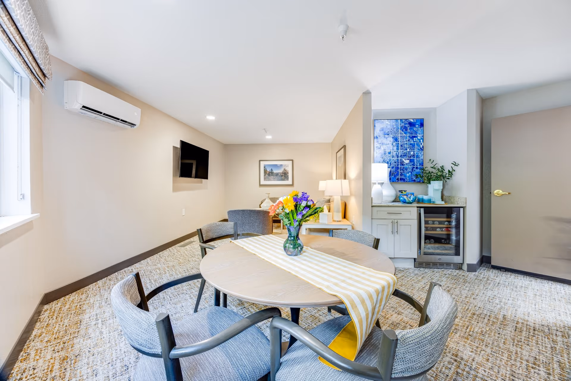A bright and cozy living area with a round wooden table in the foreground, surrounded by four cushioned chairs. A striped yellow and white table runner with a vase of colorful flowers sits on the table. In the background, there is a small sitting area with a gray armchair, a side table with a lamp, and framed artwork on the wall. To the right, a small kitchenette area features a countertop with a lamp, decorative items, a wine cooler, and a blue abstract painting above. The room has beige walls, a window with patterned curtains on the left, and a wall-mounted TV.
