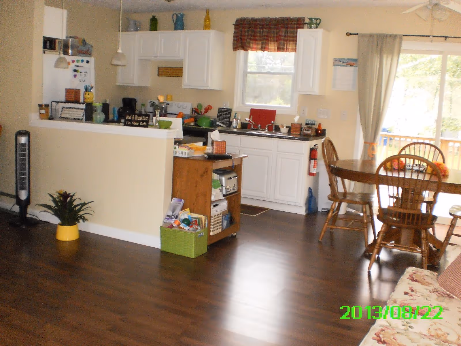 Interior view of a kitchen and dining area with wooden flooring. The kitchen has white cabinets, a stove, a sink under a window with a plaid valance, and various kitchen items on the counters. A wooden dining table with four chairs is near a sliding glass door with curtains. A small wooden shelving unit with baskets and items is next to the kitchen counter. A potted plant and a tall fan are also visible.