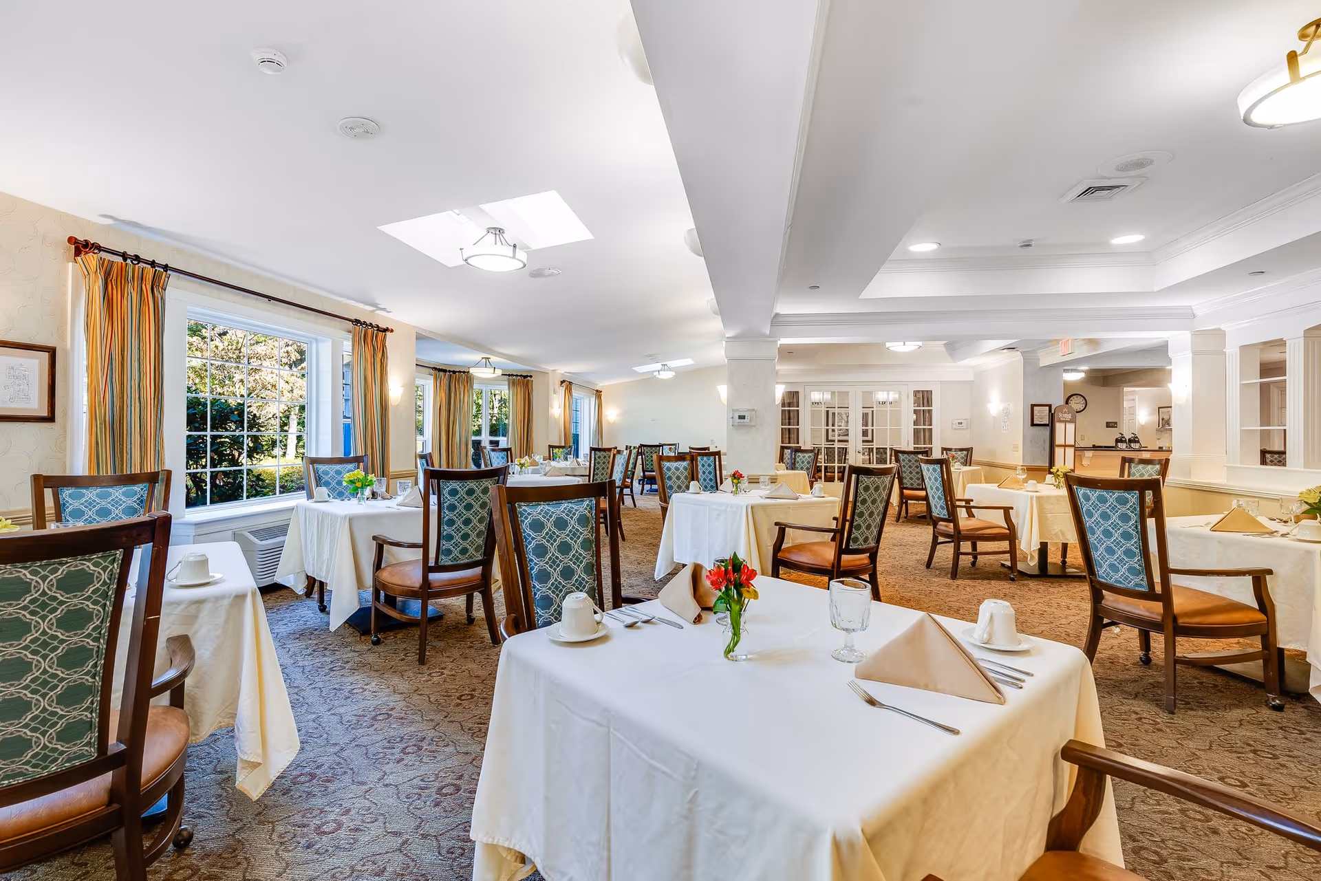 A bright and spacious dining room with multiple tables covered in white tablecloths, each set with napkins, cups, glasses, and small flower arrangements. The room features large windows with striped curtains allowing natural light to fill the space, and wooden chairs with patterned upholstery surround the tables. The carpeted floor and soft lighting create a warm and inviting atmosphere.