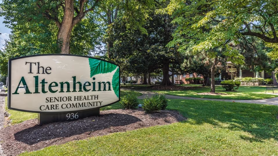 Outdoor view of The Altenheim Senior Health Care Community sign surrounded by green grass, trees, and a paved walkway leading to the building in the background.