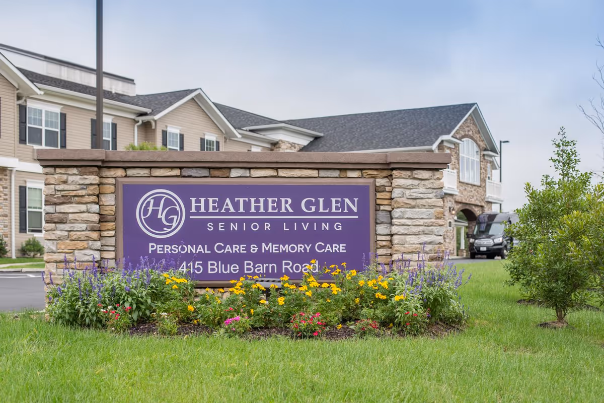 Stone and brick sign for Heather Glen Senior Living with purple background and white text, surrounded by colorful flowers and green grass, with the senior living building visible in the background under a cloudy sky.