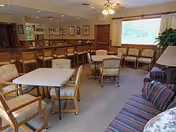 Bright senior living common room with tables, chairs, and a striped sofa near a window.