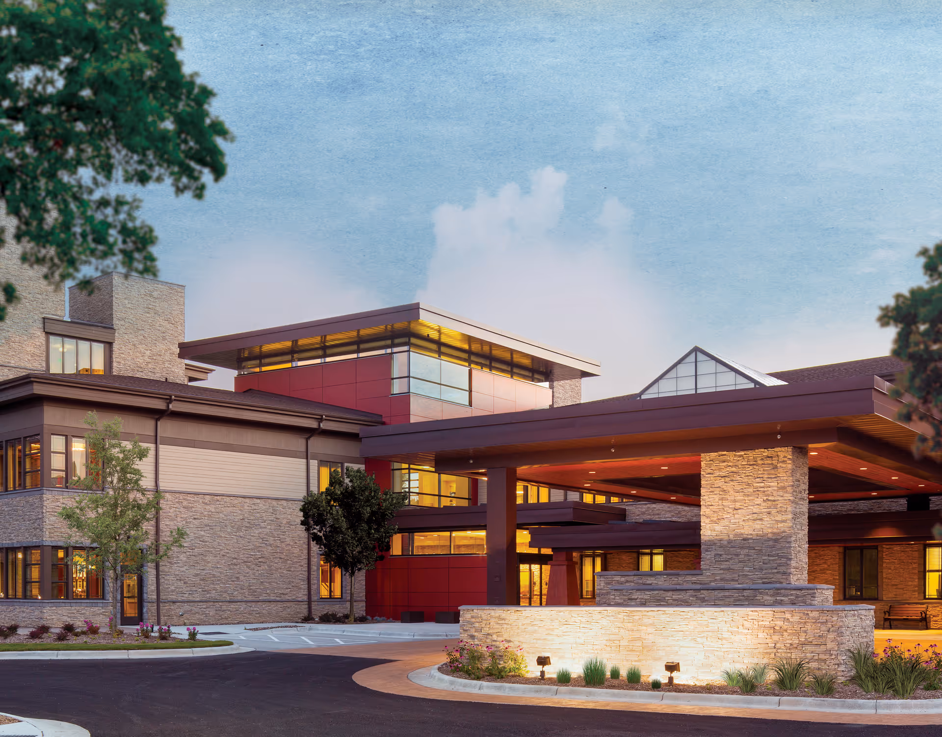 Exterior view of a modern senior living facility building with stone and red panel accents, large windows, and a covered entrance with stone pillars. The sky is clear with some clouds, and there are trees and landscaping around the building.