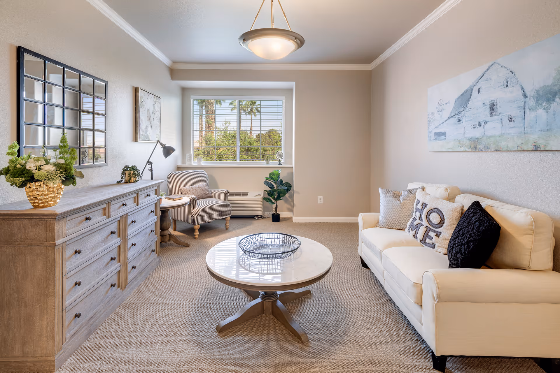 A bright and cozy living room with a beige sofa adorned with decorative pillows, including one with the word 'HOME'. A round wooden coffee table with a decorative tray sits in the center on a carpeted floor. To the left, there is a wooden dresser with a gold vase holding green and white flowers, a small lamp, and a striped armchair next to a window with blinds. A large window shows palm trees and greenery outside. The walls are painted light beige with crown molding, and a large painting of a barn hangs above the sofa.