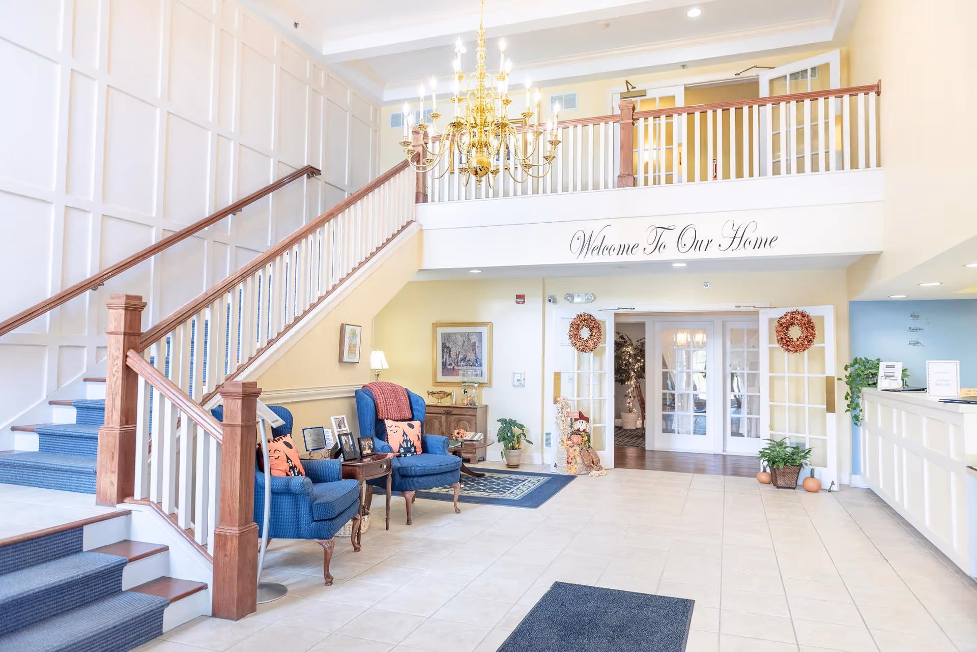 Bright and welcoming senior living facility lobby with a staircase on the left, two blue armchairs with orange pillows, a chandelier hanging from the ceiling, and a reception desk on the right. The wall above the entrance reads 'Welcome To Our Home'.