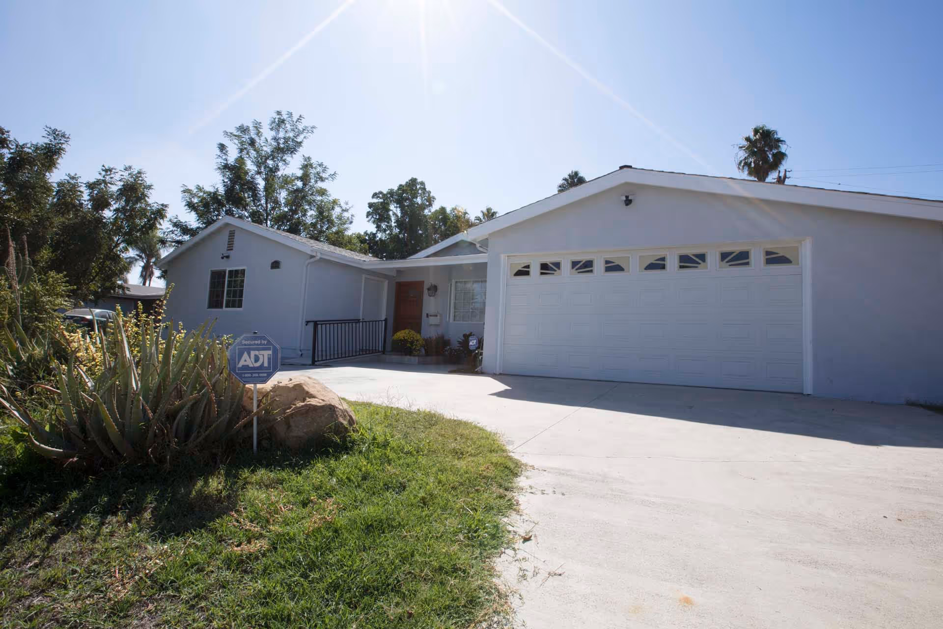 Front exterior view of a single-story residential building with a white garage door, a driveway, and a small garden area with plants and a security sign in front.