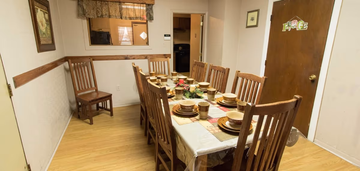 A dining room with a long wooden table set for eight people with matching wooden chairs. The table is covered with a tablecloth and has plates, bowls, cups, and a centerpiece. The room has light-colored walls, a wooden floor, a window with a valance, and a closed wooden door with a decorative sign that says 'Peace'.