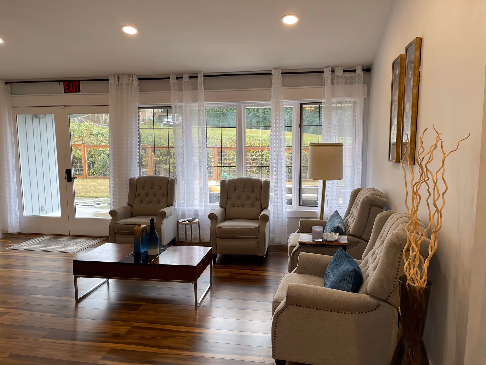 A cozy living room area with four beige upholstered armchairs arranged around a wooden coffee table. The room has large windows with sheer white curtains allowing natural light to brighten the space. There is a floor lamp beside one of the chairs and decorative items including blue cushions, vases on the coffee table, and tall dried branches in a vase near the wall. The floor is wooden, and a glass door leads outside to a fenced garden area.