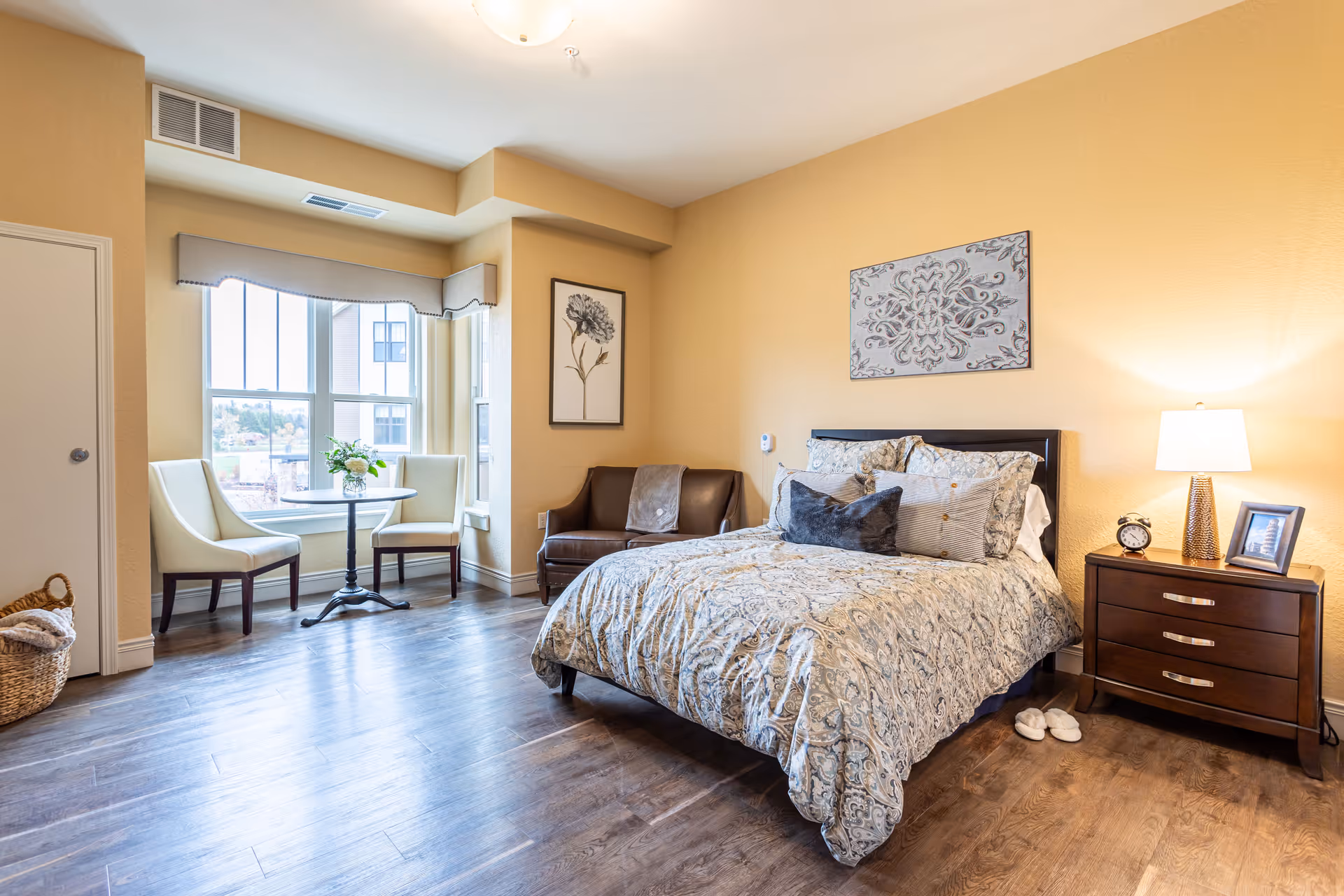 A cozy bedroom with a large bed covered in patterned bedding and multiple pillows. Next to the bed is a wooden nightstand with a lamp, alarm clock, and framed photo. There is a small brown loveseat against the wall and a round table with two white chairs near a large window letting in natural light. The walls are painted a warm beige color, and there are two pieces of framed artwork hanging on the walls. The floor is a dark wood laminate.