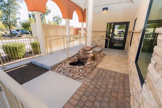 Entrance area of a senior living facility with a ramp, rock water feature, arched columns and glass doors.