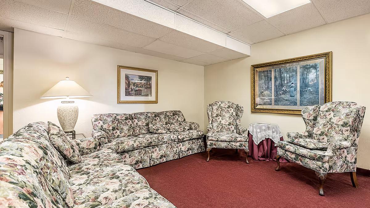 A cozy sitting area with floral patterned sofas and armchairs arranged on a red carpet. There is a side table with a white lace cloth and a lamp on another side table. Two framed paintings hang on the beige walls under a white drop ceiling with fluorescent lighting.