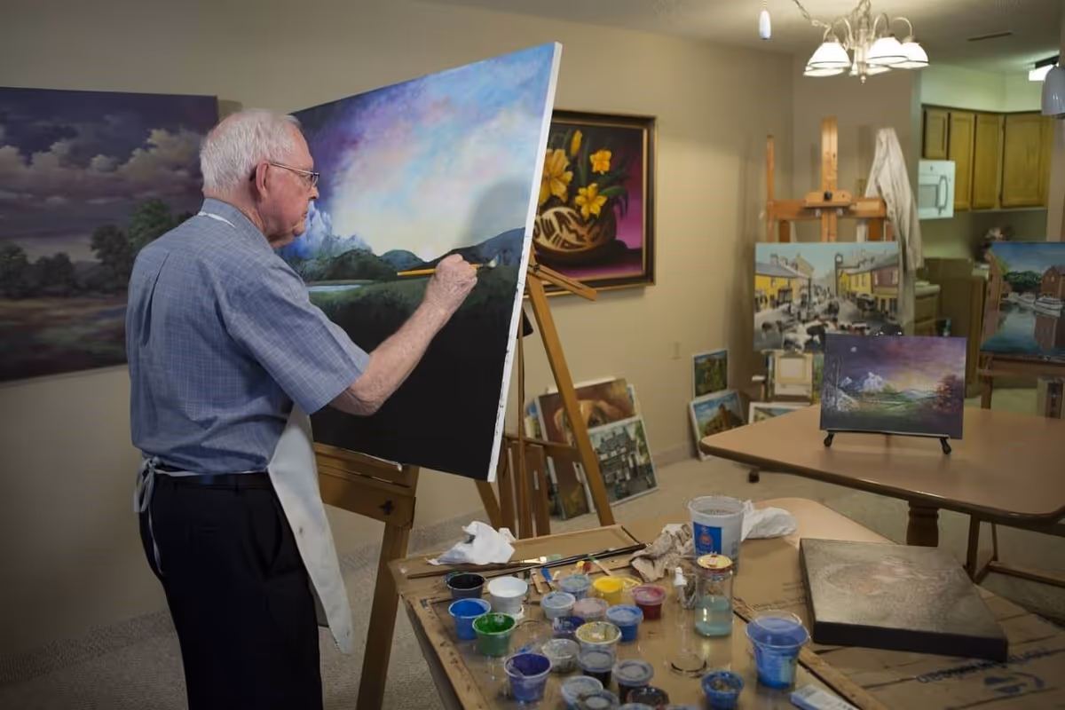 An elderly man wearing glasses and a blue shirt is painting a landscape on a canvas in an art studio. The room contains several easels with paintings, a table with various paint containers and brushes, and a kitchen area in the background.