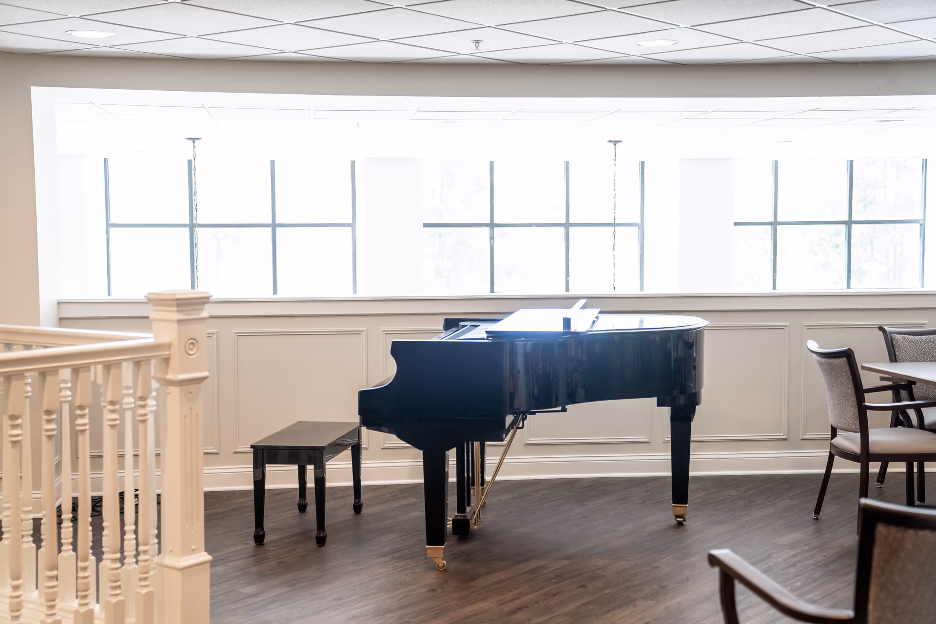A bright room with large windows featuring a black grand piano and matching bench on a wooden floor. To the right, there are chairs and a table, and to the left, a white railing is visible.
