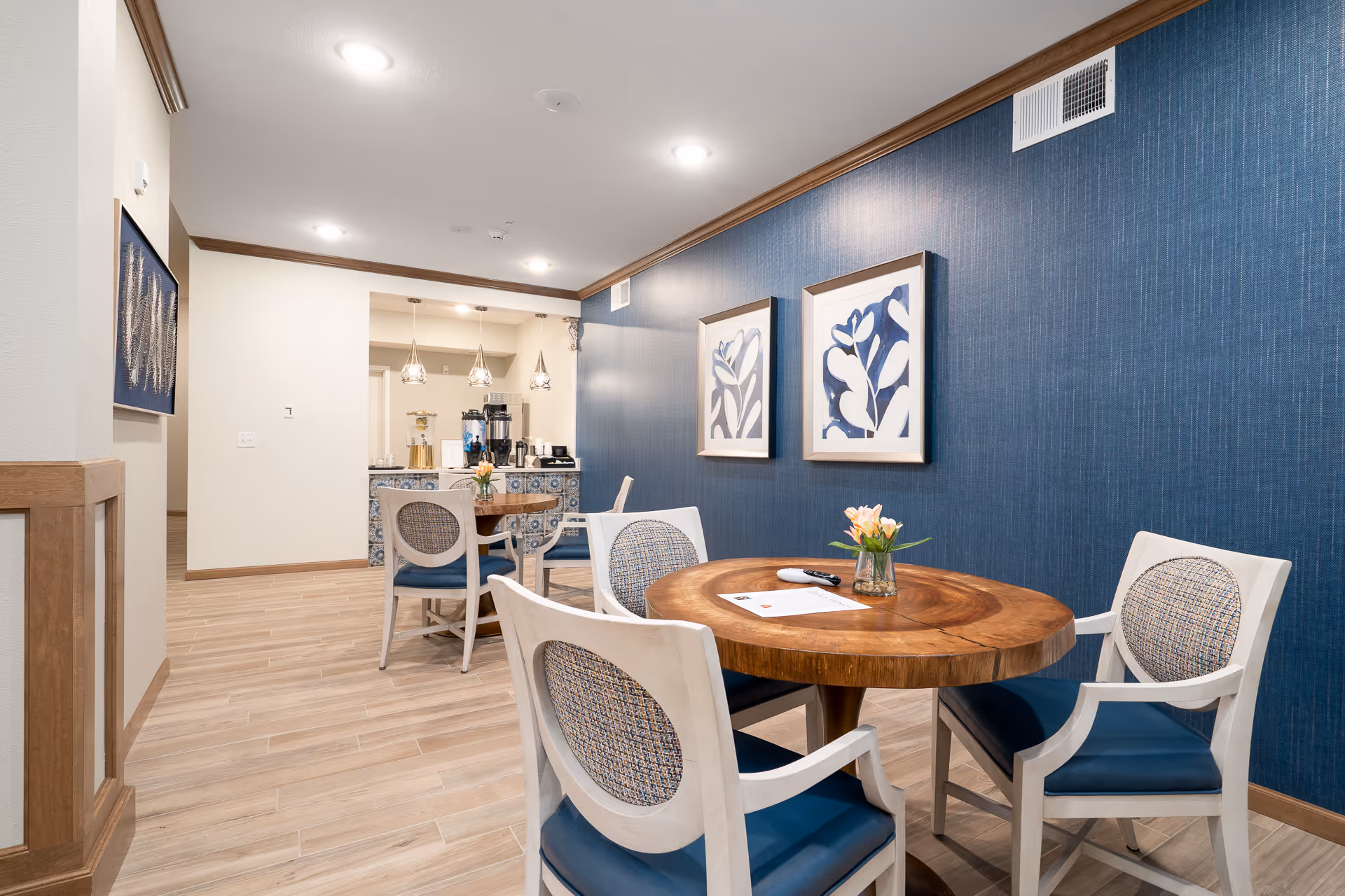 Round wooden table with four upholstered chairs in a well-lit dining area featuring a blue accent wall, framed artwork, and a coffee station in the background.