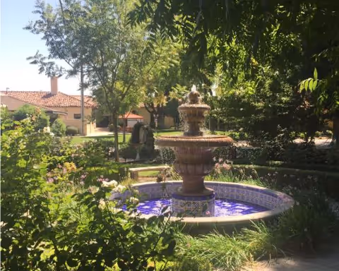 A peaceful outdoor garden area with a decorative multi-tiered fountain surrounded by greenery, flowers, and trees. In the background, there is a building with a red-tiled roof partially visible under the sunlight.