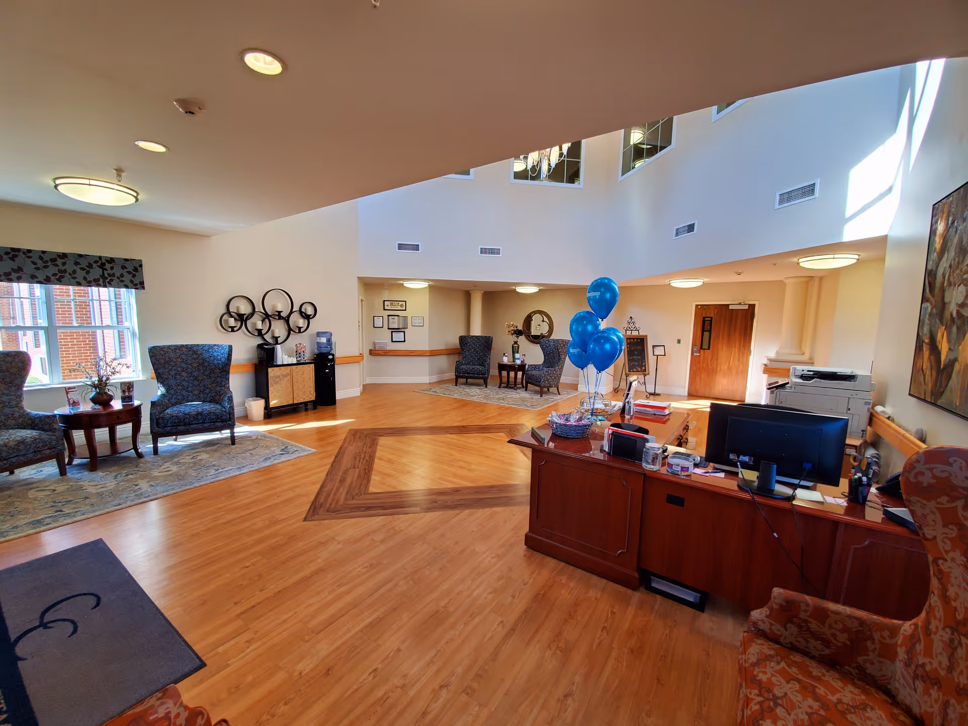 A spacious and well-lit reception area of Carrington Cottage Memory Care with wooden flooring and a high ceiling. The room features a wooden reception desk with a computer, blue balloons, and office supplies. There are several upholstered chairs and small tables arranged on area rugs near windows and walls. Decorative wall art and a water dispenser are also visible.