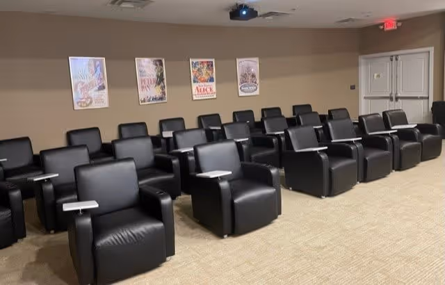 A small theater room with three rows of black leather recliner chairs, each equipped with a small attached table. The walls are beige and decorated with framed posters of classic movies. A ceiling-mounted projector is visible, and there is a double door at the back of the room.