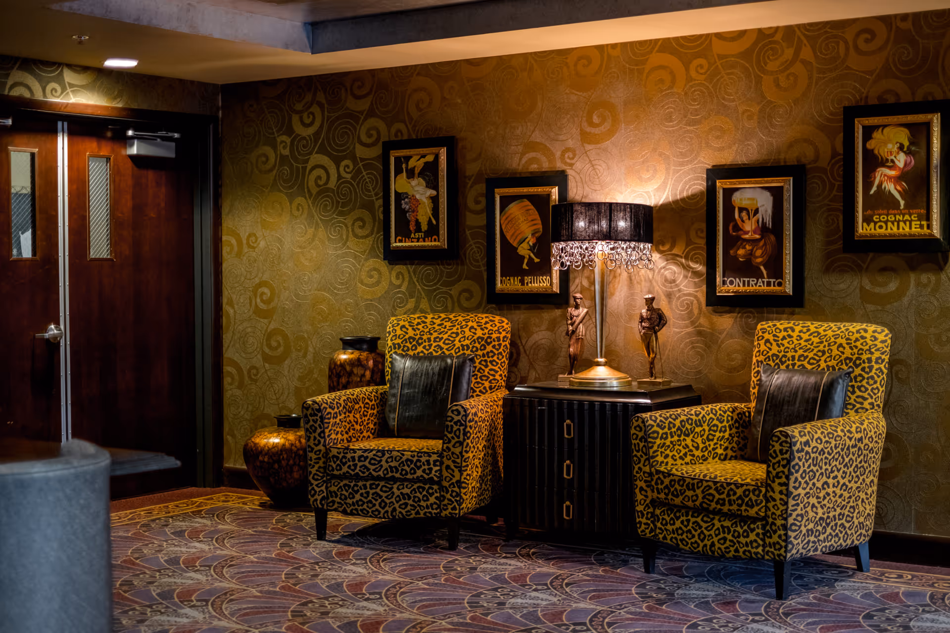 A cozy interior seating area with two leopard print armchairs, each with a black cushion. Between the chairs is a small black cabinet with a decorative lamp and two statuettes on top. The wall behind features a gold patterned wallpaper and four framed vintage-style posters. To the left, there is a set of double wooden doors with small windows.
