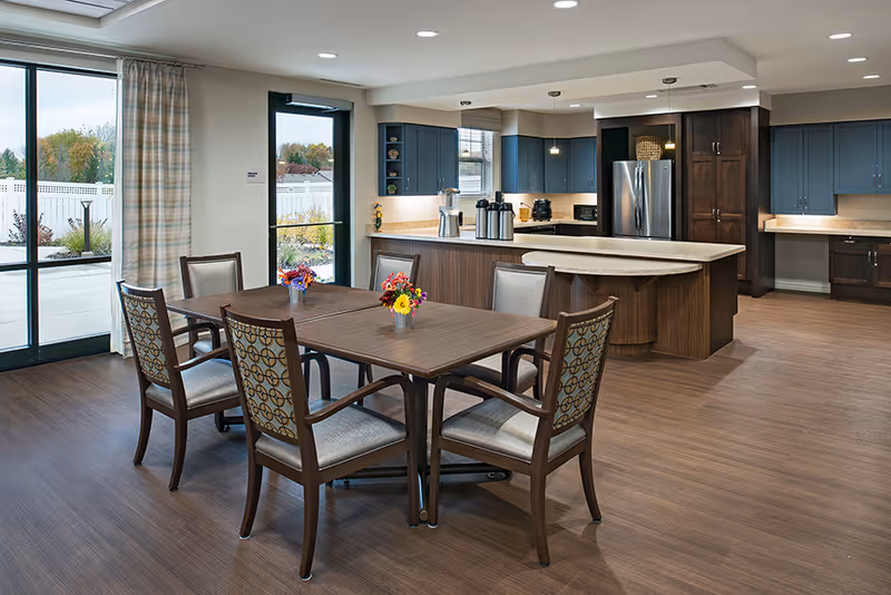 A spacious kitchen and dining area with a wooden dining table surrounded by six chairs. The kitchen features dark blue cabinets, a large island with a light countertop, stainless steel refrigerator, and various kitchen appliances. Large windows and a glass door provide natural light and a view of an outdoor patio area.