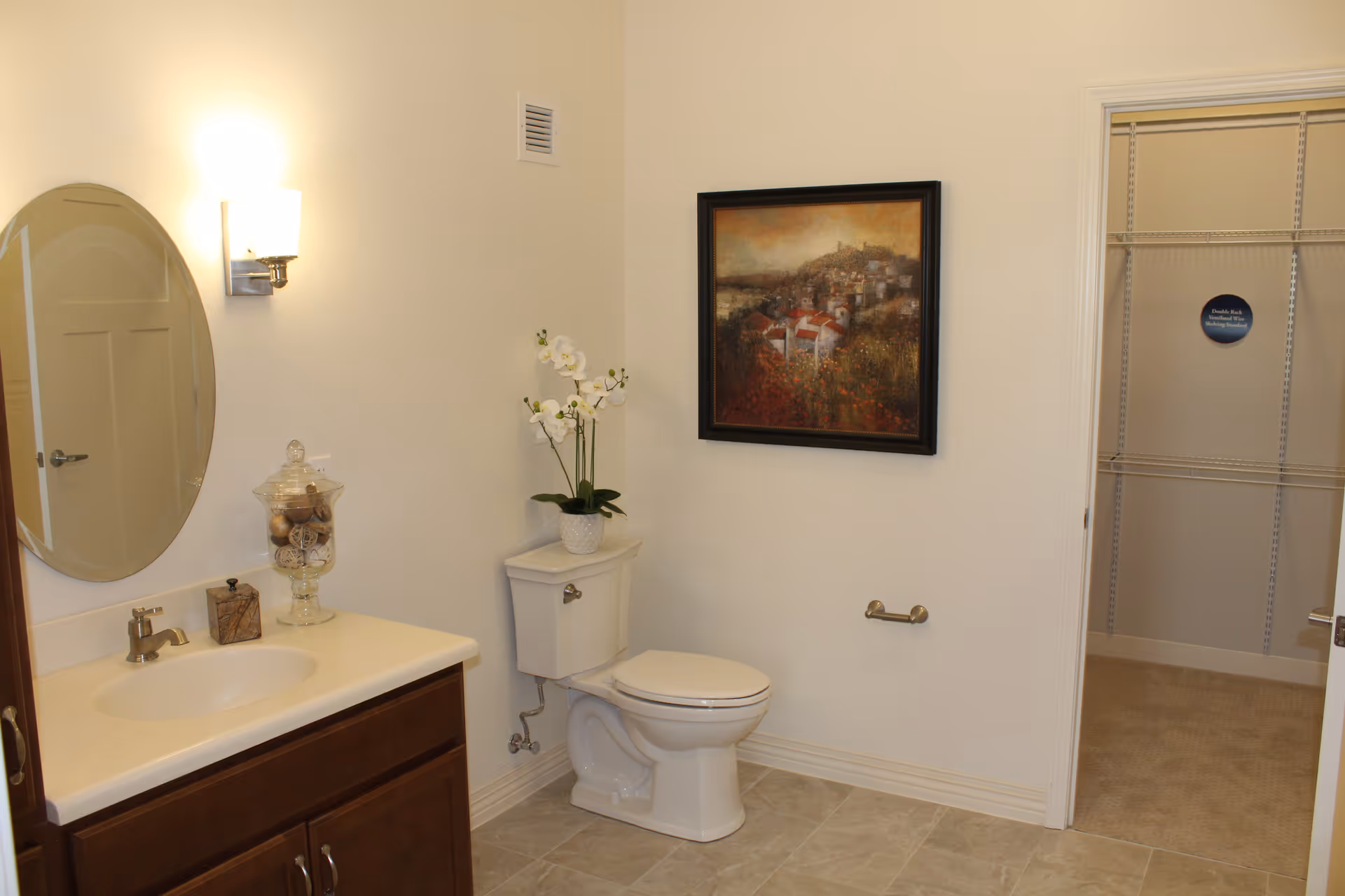 A clean bathroom with a white toilet and a wooden vanity with a sink. Above the vanity is an oval mirror and a wall-mounted light fixture. A decorative glass jar and a small box are on the vanity. A potted orchid plant is placed on the toilet tank. A framed painting of a landscape hangs on the wall above the toilet. An open door reveals a walk-in closet with shelving.