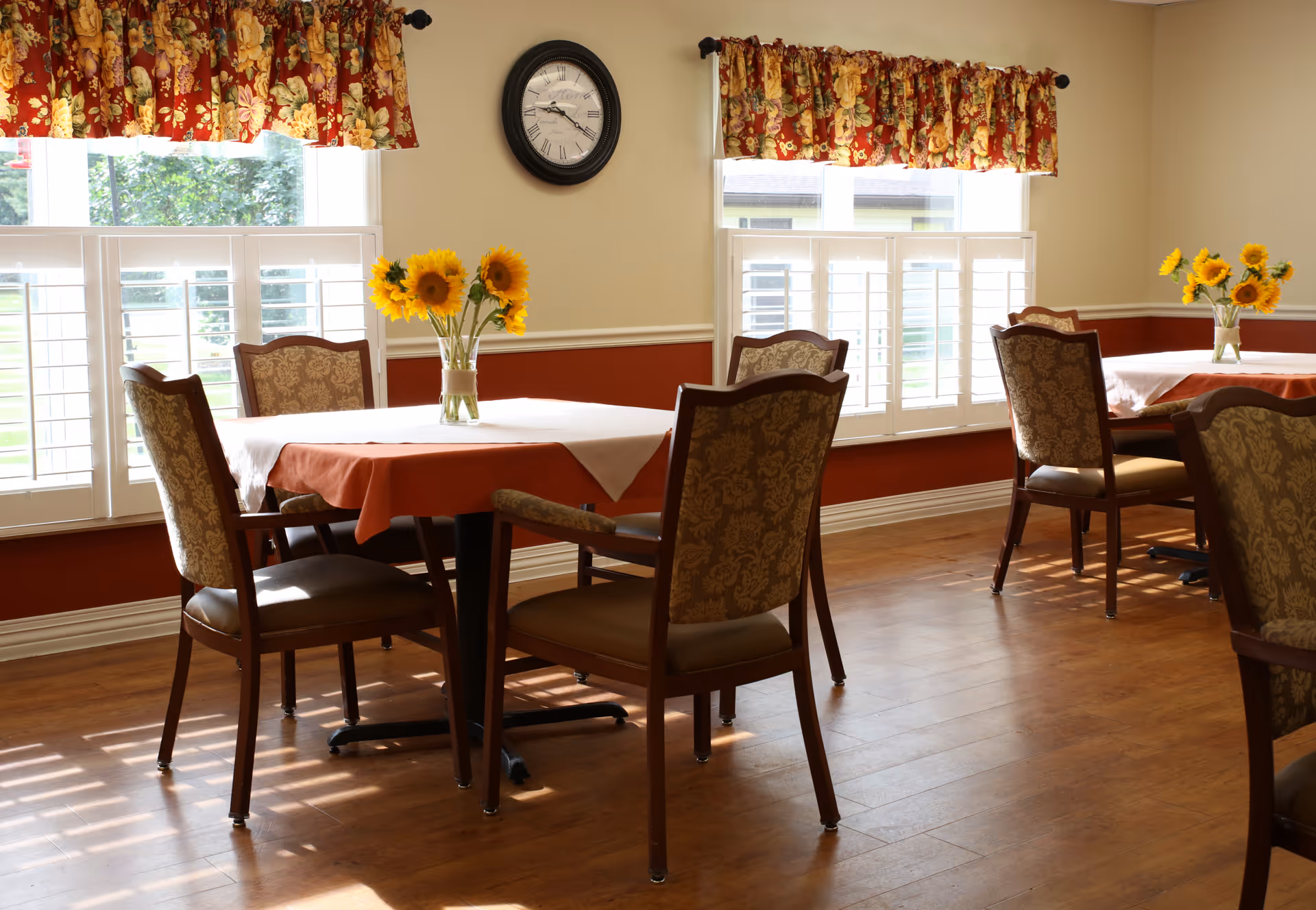 A bright dining room with wooden floors, two tables covered with white and orange tablecloths, each adorned with a vase of sunflowers. The room has large windows with white shutters and floral valances, and a round wall clock is mounted on the beige wall.