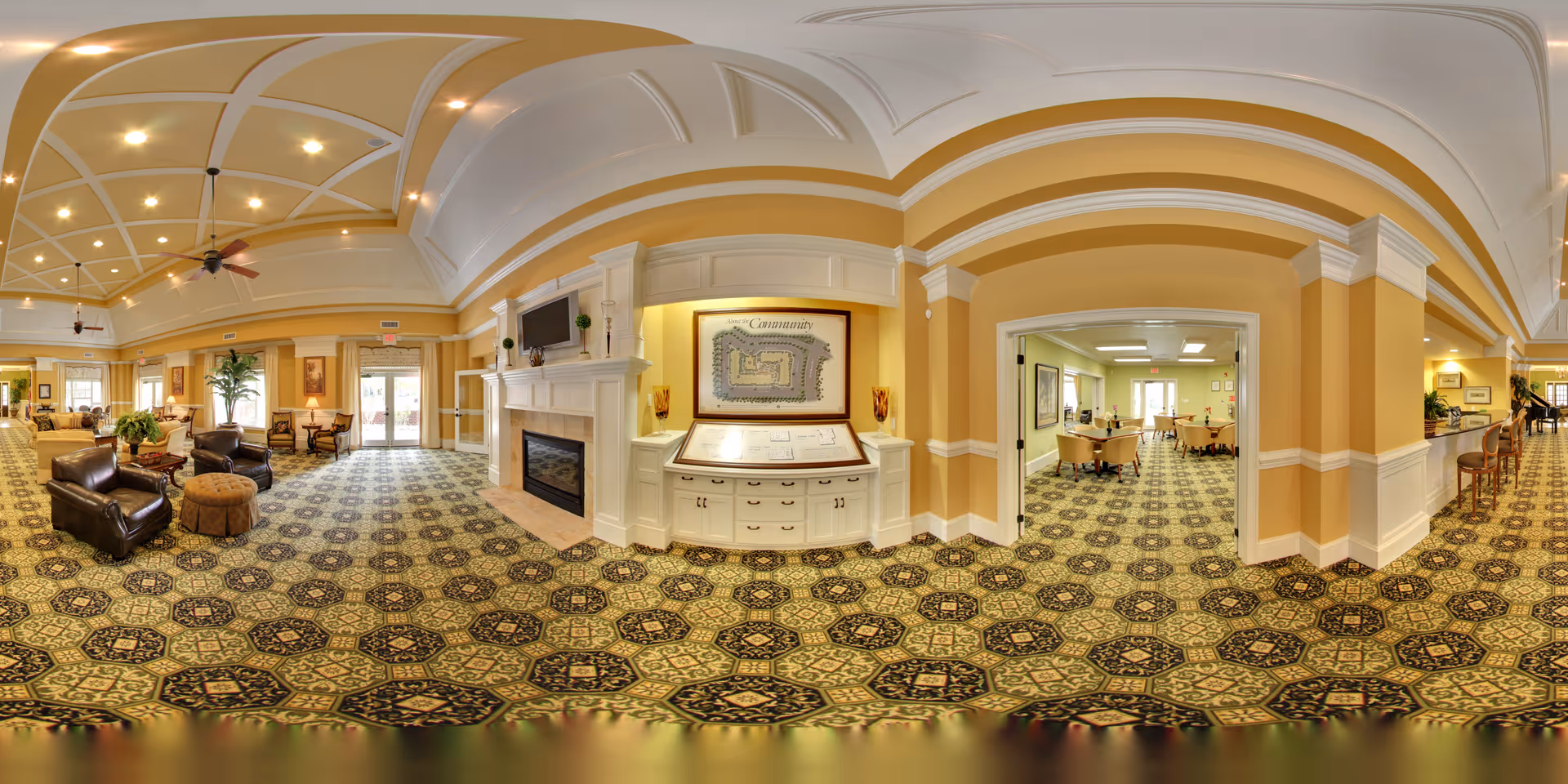 A spacious and well-lit senior living facility common area with patterned carpet, beige walls, and a white coffered ceiling with recessed lighting and ceiling fans. The room features comfortable leather armchairs, a fireplace with a TV mounted above it, and a framed community map on the wall. To the right, there is an open doorway leading to a dining area with tables and chairs. The space is decorated with plants and artwork, creating a warm and inviting atmosphere.