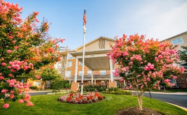 Exterior view of Arbor Terrace Herndon with flowering trees and a flagpole at the entrance
