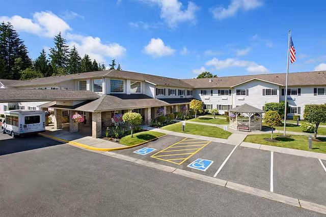 Exterior view of The Bridge at Mount Vernon senior living facility showing a two-story building with a covered entrance, a small gazebo in a landscaped courtyard, handicap parking spaces, and an American flag on a flagpole under a blue sky with some clouds.