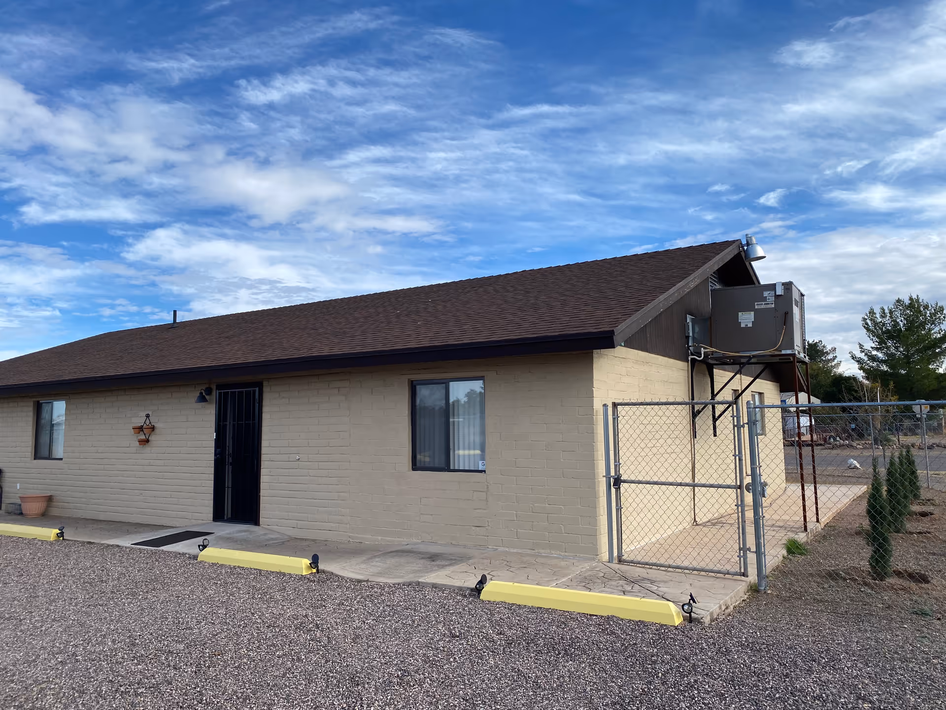Single-story beige brick building with a brown roof, a black metal door, windows, a chain-link side gate and an exterior HVAC unit under a partly cloudy sky.