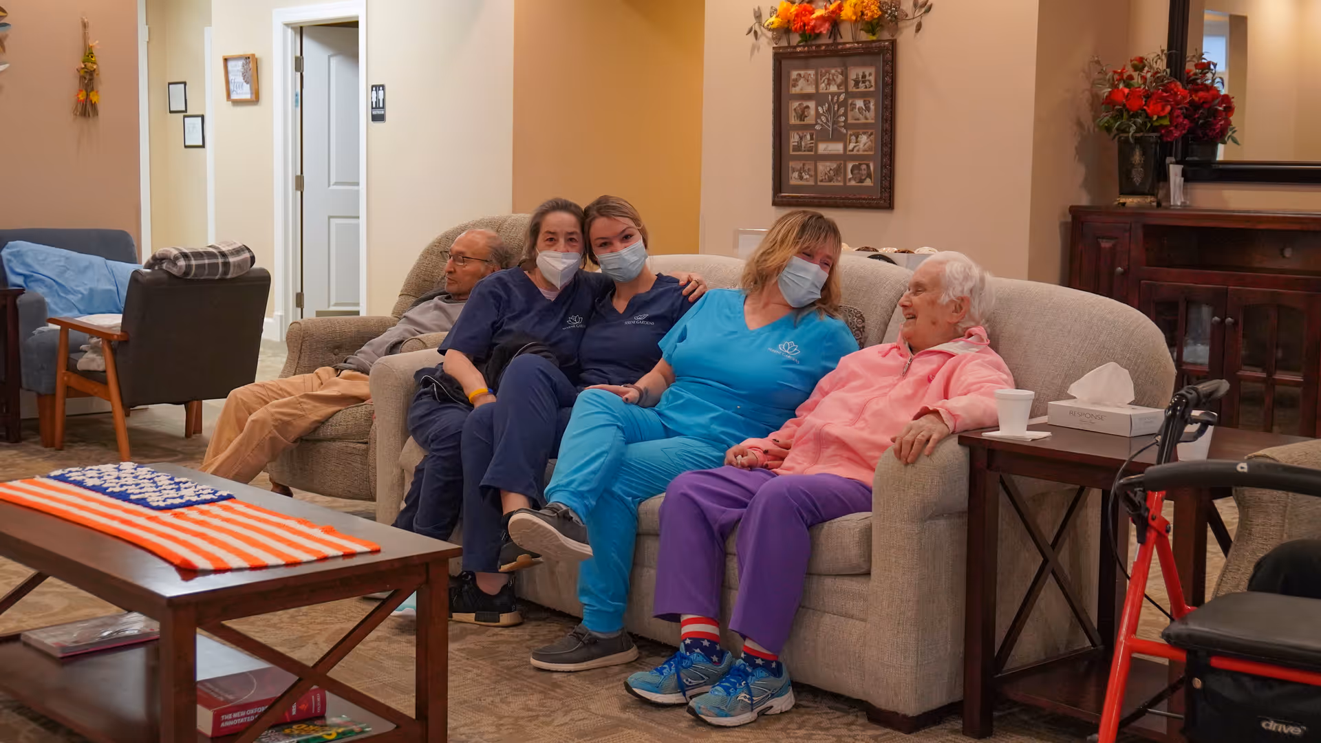 A group of four people sitting on a beige couch in a cozy living room. Two elderly individuals and two caregivers wearing scrubs and face masks are seated together, smiling and interacting. The room has warm lighting, a wooden coffee table with a knitted American flag on it, a side table with a tissue box and a cup, and a walker nearby. The background shows a hallway and some framed pictures on the wall.