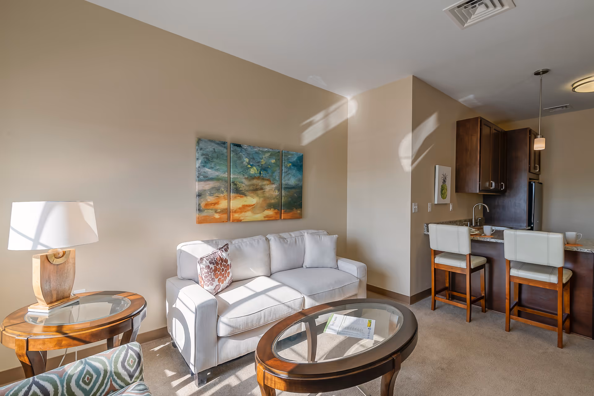 Sunlit living room with a white sofa, glass-topped tables, wall art, and a small kitchen bar with two stools.